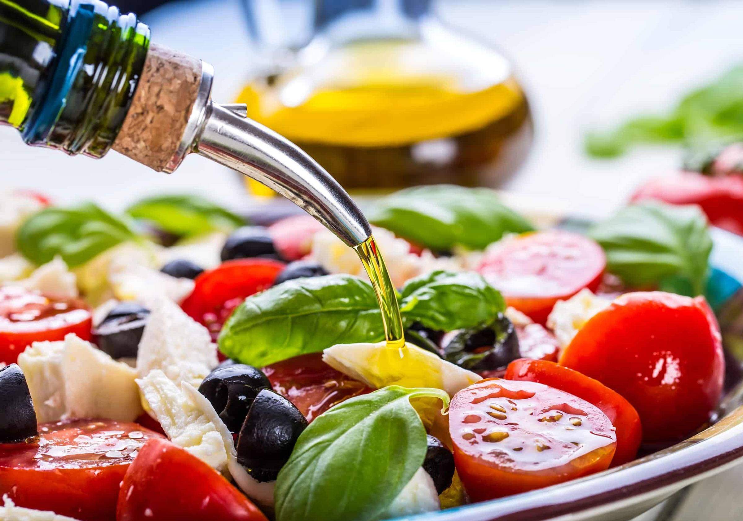 Olive oil being poured over a fresh salad with basil, cherry tomatoes, black olives, and cheese. A glass bottle of olive oil is visible in the background.