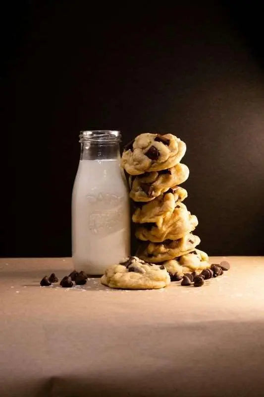 Chocolate chip cookies stacked beside glass milk bottle with scattered chocolate chips