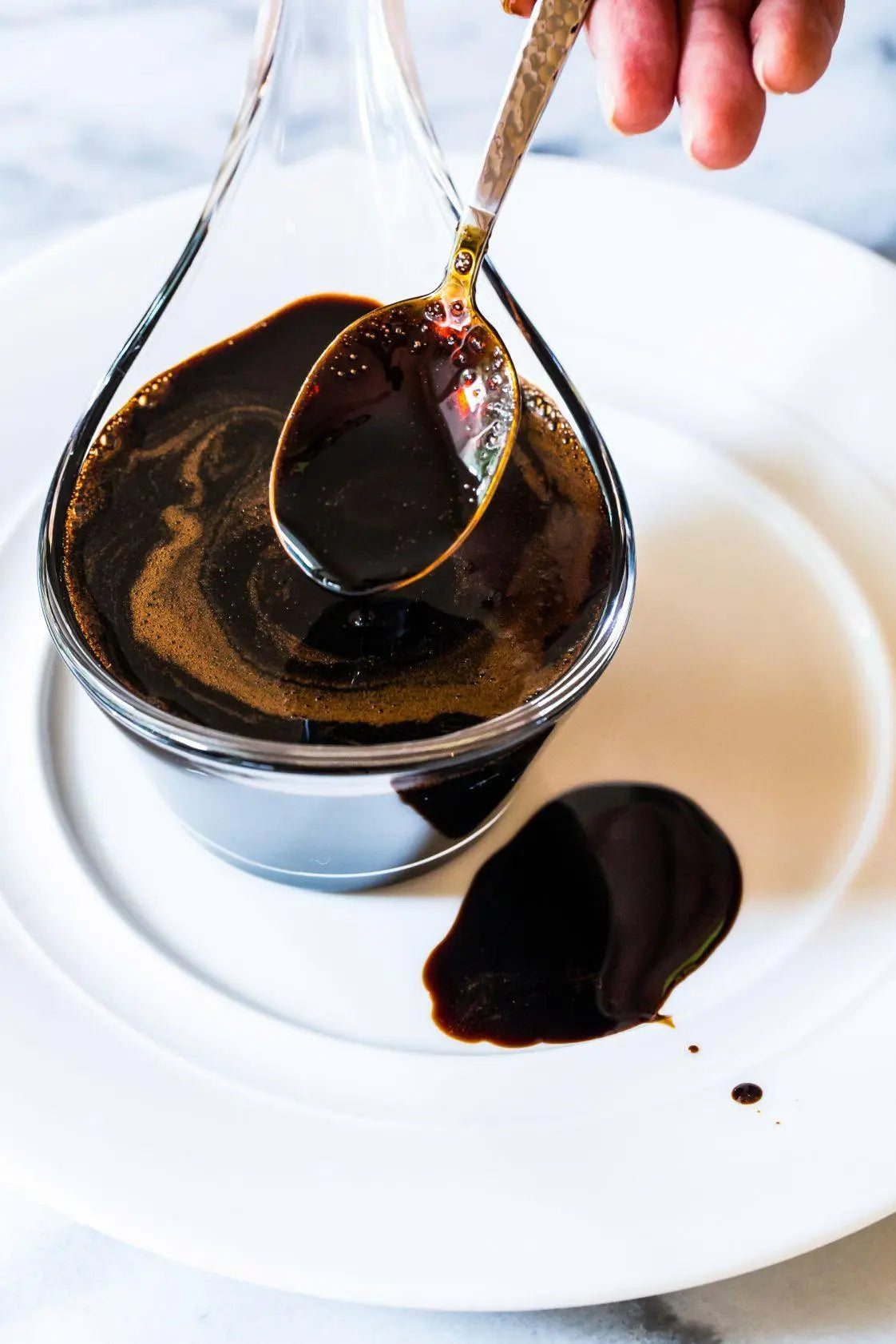 Rich balsamic vinegar drizzled from spoon over white plate, showcasing artisanal quality for gourmet cooking.