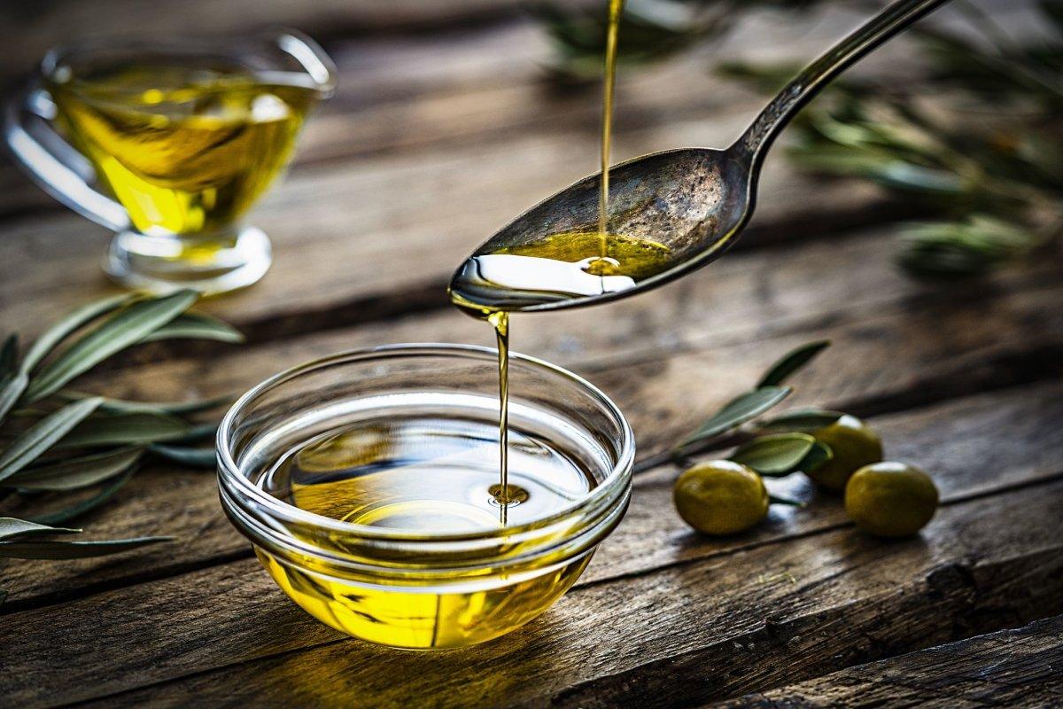 Golden olive oil drizzling from spoon into glass bowl, fresh olives & leaves on rustic wood