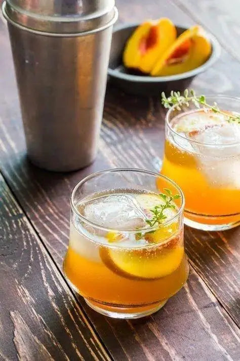 Peach cocktail with thyme garnish, served in glasses with ice on rustic wood table