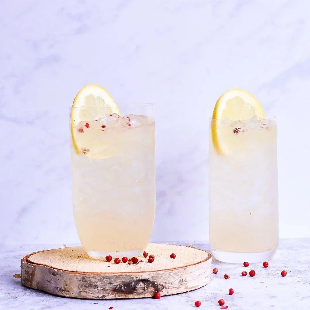 Two glasses of lemon-infused cocktail with pink peppercorns, served on a birch wood slice, perfect for refreshing summer drinks.