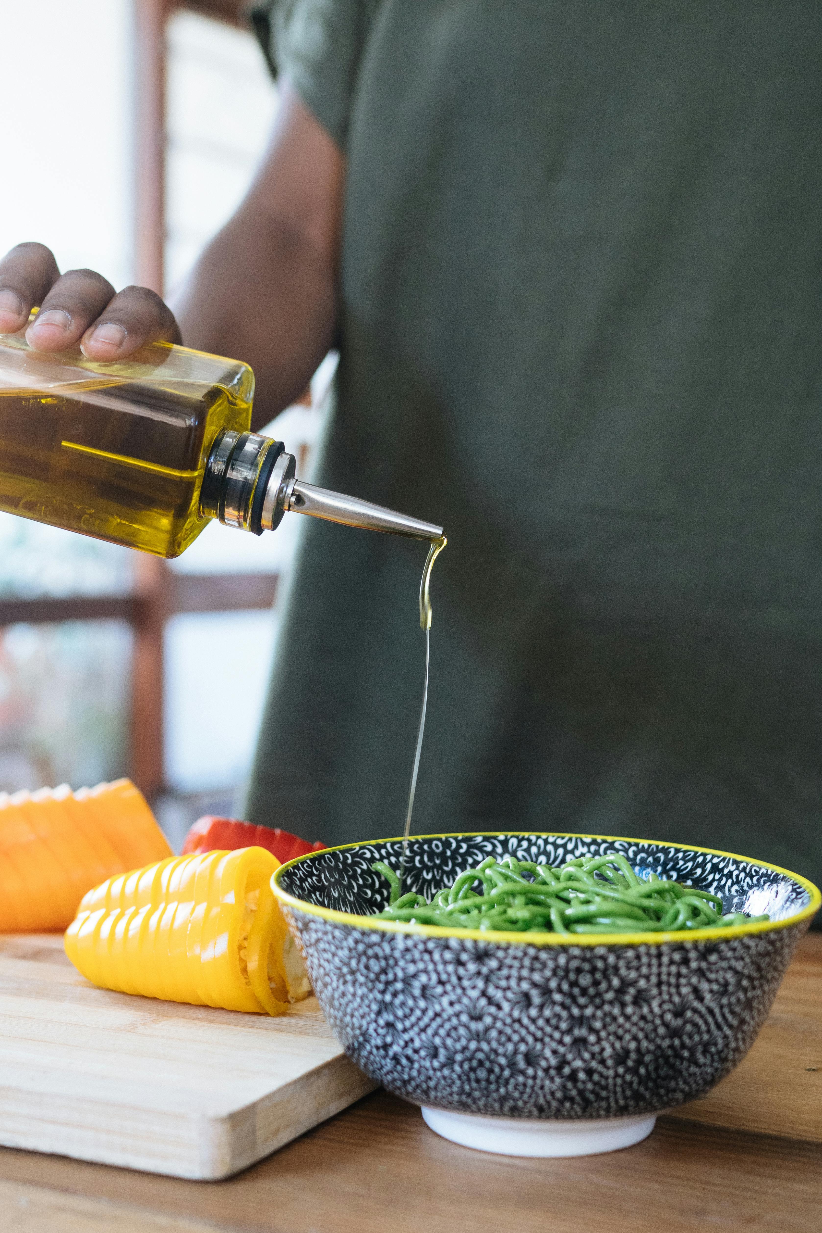 Drizzling premium olive oil over green noodles with fresh peppers, perfect for gourmet cooking.