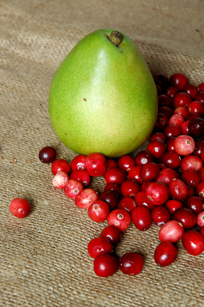 A green pear rests on a textured fabric beside a scattered pile of red cranberries, capturing the fruity crisp essence of Tastefully Olive's Cranberry Pear White Balsamic Vinegar.
