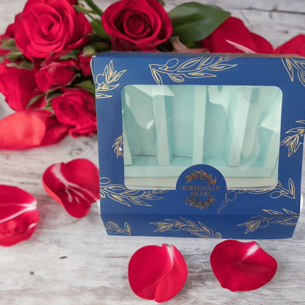 A blue cardboard box with gold decorations and a transparent front window, labeled as the "Luxurious 60ml Gift Box" from Tastefully Olive, sits on a white surface surrounded by red rose petals and a bouquet of red roses, presenting a sampler set of luxurious flavors.