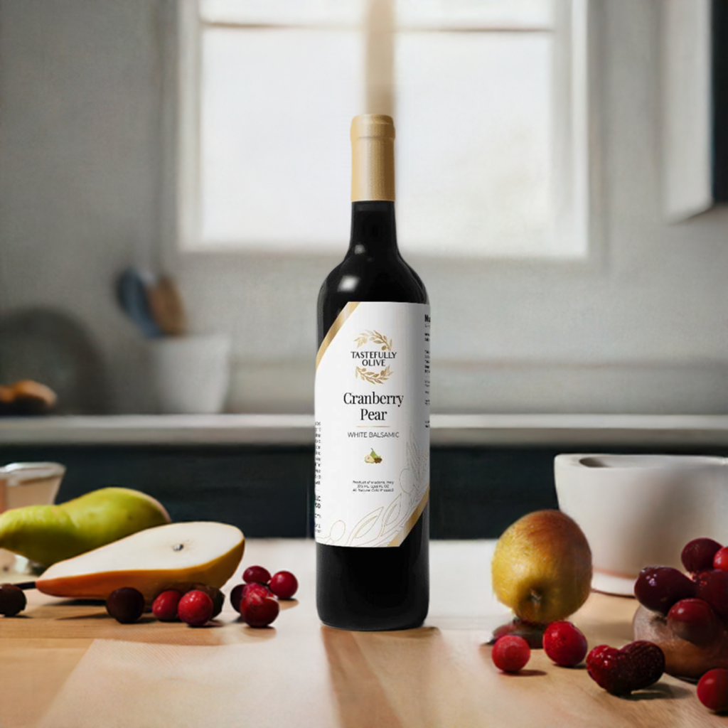 A bottle of Tastefully Olive's Cranberry Pear White Balsamic Vinegar sits on a kitchen countertop, encircled by cranberries, pears, a mortar and pestle, and various kitchen items. The fruity, crisp taste of this vinegar pairs perfectly with flavored olive oils for a delightful culinary experience.