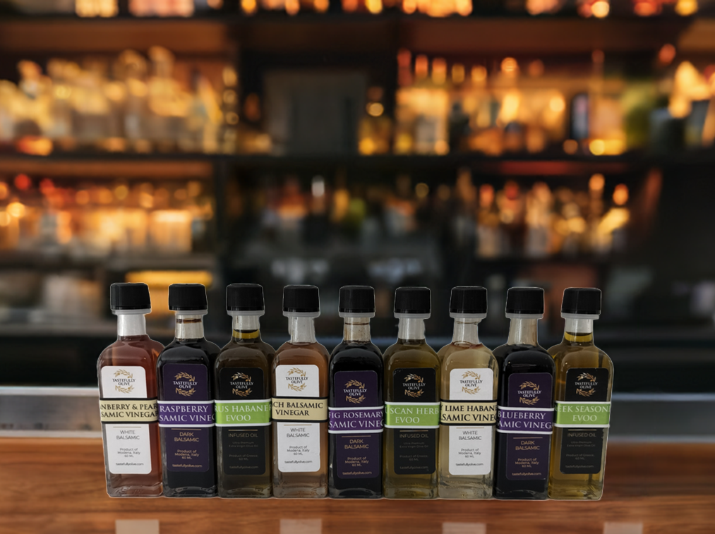 A variety of Tastefully Olive Individually Sized 60ml bottles are lined up on a wooden bar counter in front of a blurred background of shelves stocked with assorted bottles.