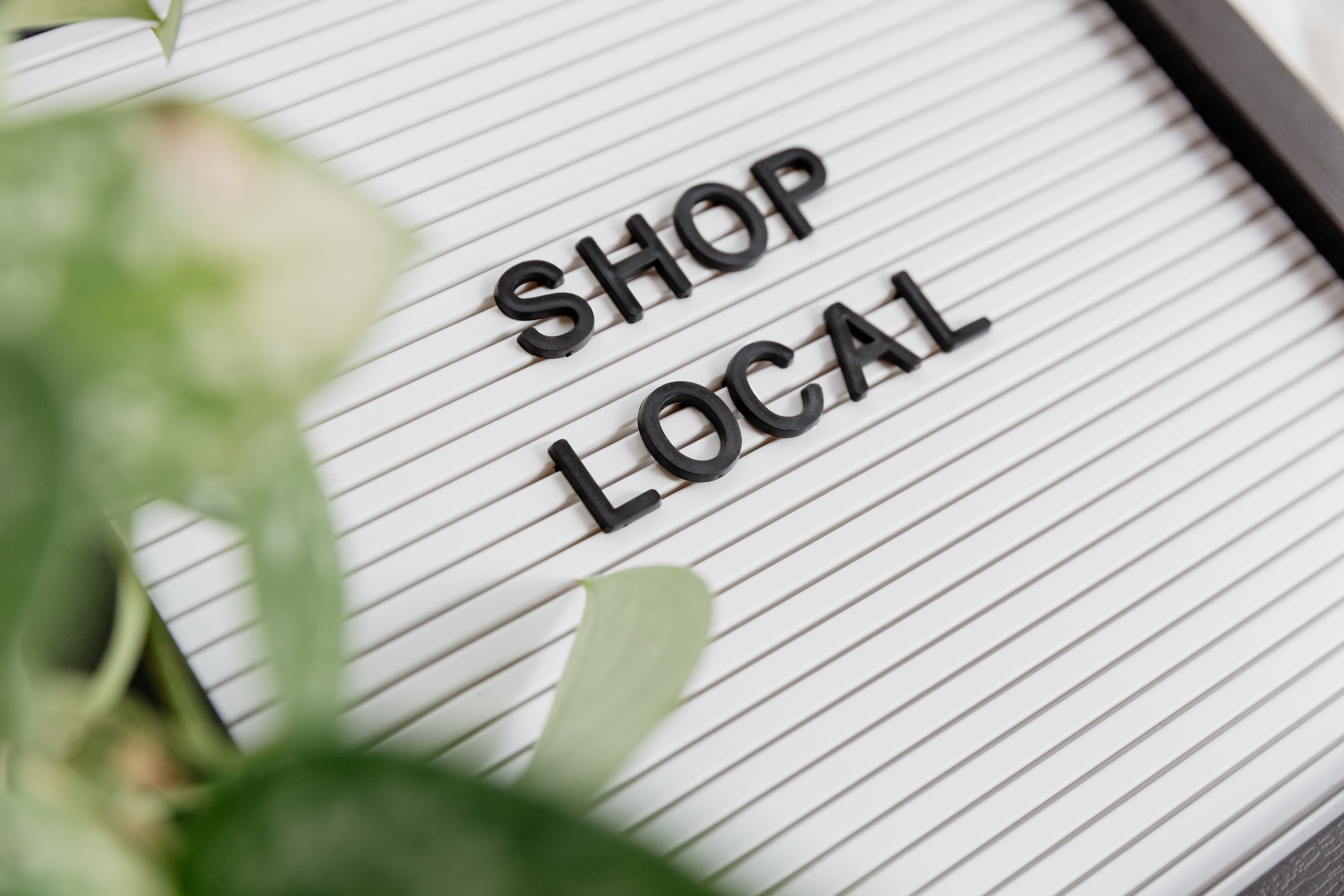 A signboard with a white, ribbed background features the black, uppercase text "SHOP LOCAL." Green plant leaves partially frame the corners of the image, adding a natural touch to the scene. The image emphasizes the importance of supporting local businesses.