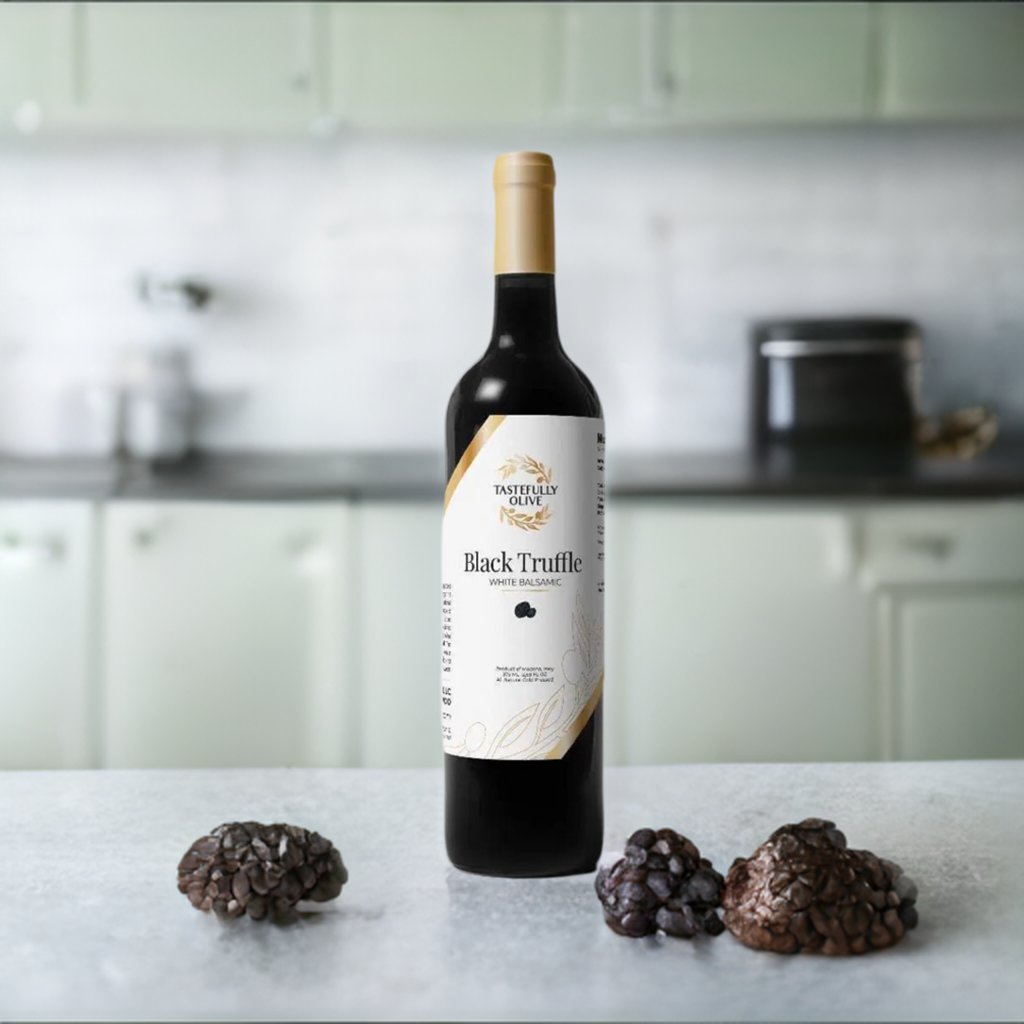 A bottle of Tastefully Olive Black Truffle White Balsamic Vinegar stands on a kitchen counter, flanked by black truffles on both sides. The background features light-colored cabinets and kitchen utensils, perfect for creating delicious pasta recipes that highlight the umami flavor of black truffles.