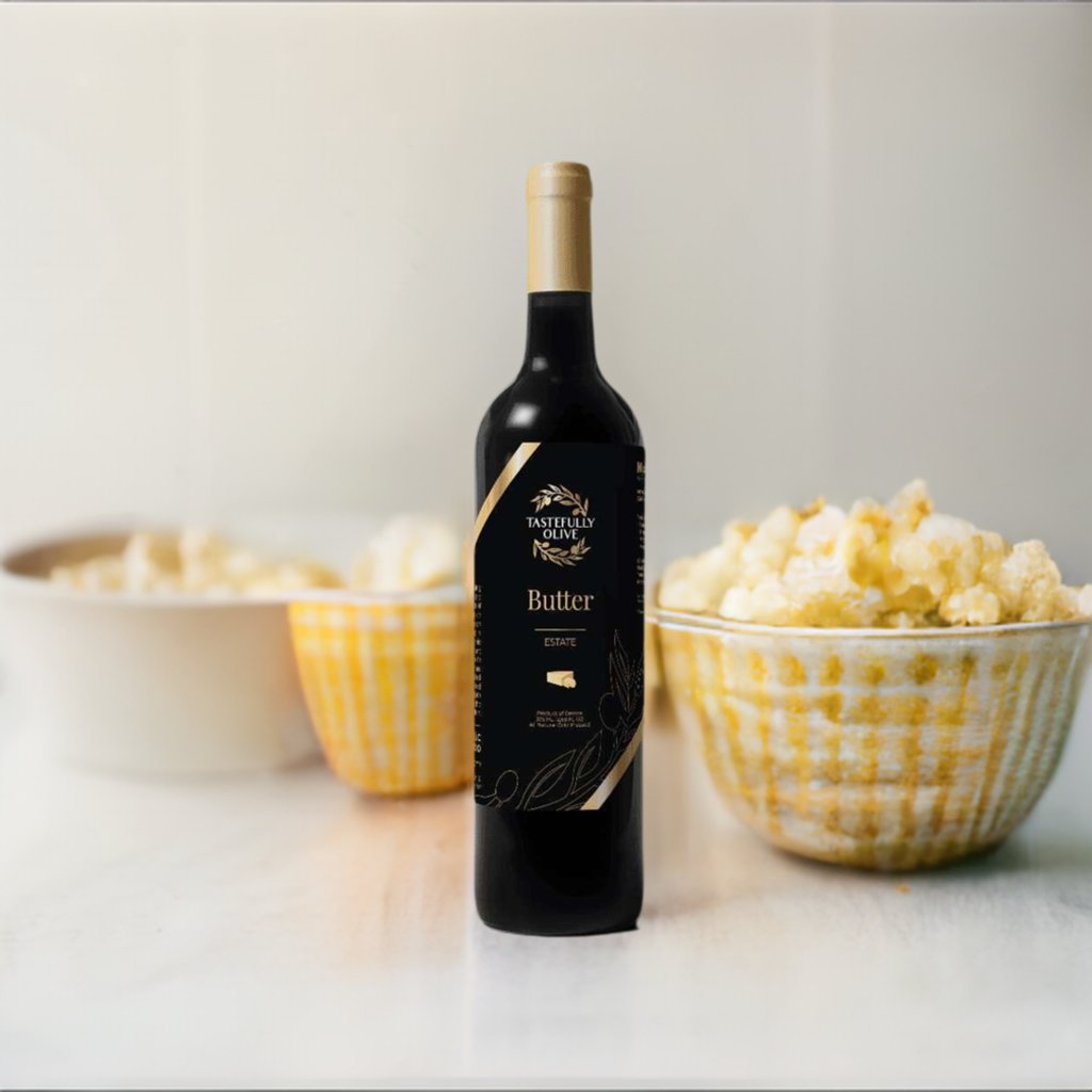 A bottle of Tastefully Olive Butter Infused Olive Oil stands on a white surface, flanked by two bowls of popcorn, delivering a savory buttery taste perfect for a cozy movie night.