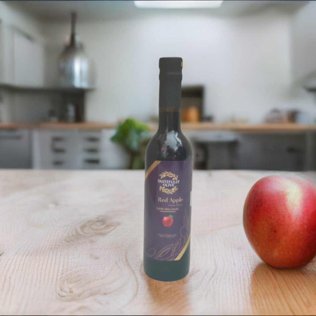 A bottle of Tastefully Olive's Red Apple Dark Balsamic Vinegar and a red apple placed on a wooden kitchen countertop, perfect for dressings and dessert sauces, with a blurred kitchen background.